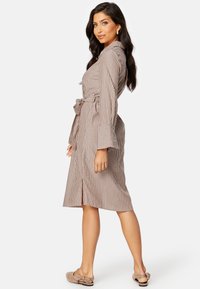 Robe a righe marrone con vita fasciata, maniche svasate e taglio al ginocchio, realizzata in un tessuto leggero. Indossata con scarpe beige senza lacci.