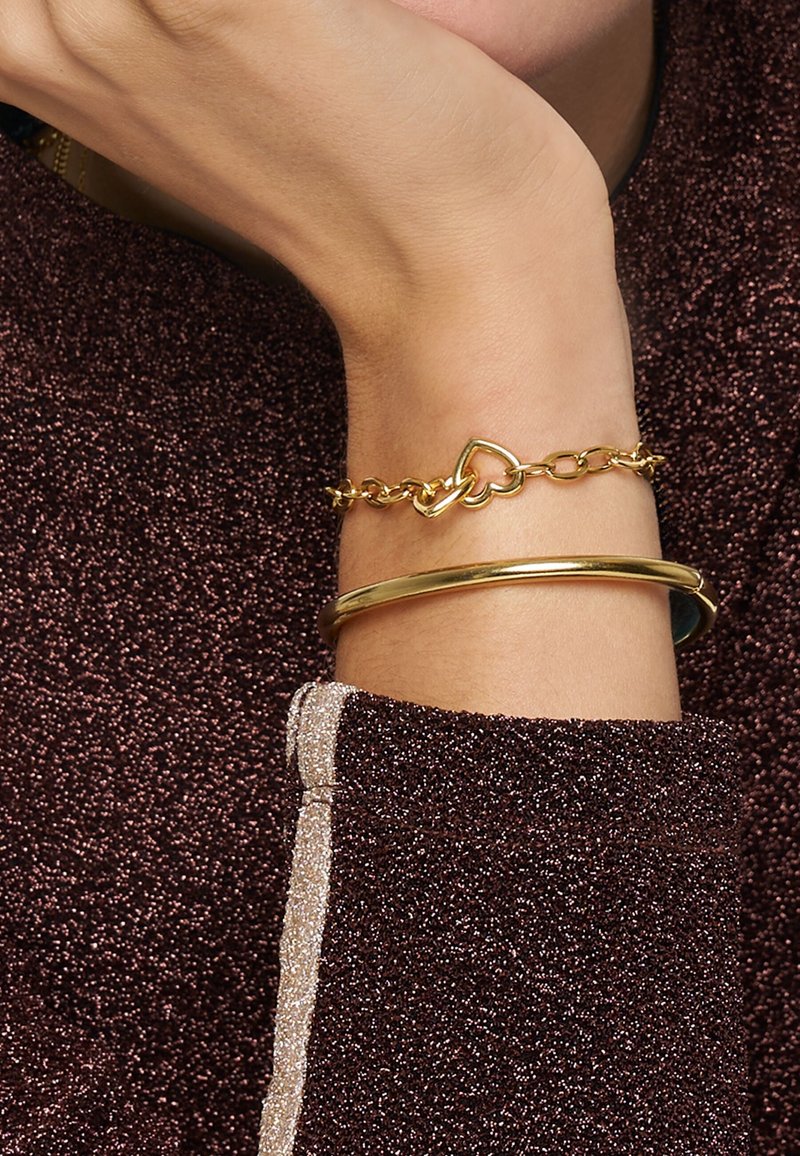 Pulseras de oro: una unida con formas de corazón, la otra es un brazalete liso. Mano de tono de piel descansando sobre un tejido oscuro y texturizado.