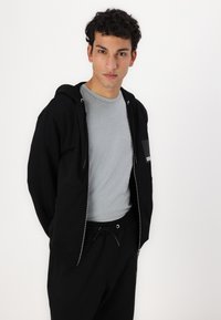 Sort hættejakke med lynlås og sidelommer, båret over en lysegrå t-shirt, parret med sorte sweatpants med snoretræk. Enkel, afslappet design.