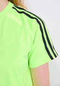 Νεον πράσινη αθλητική μπλούζα με μαύρες ρίγες κατά μήκος των ώμων. Φτιαγμένη από λείο ύφασμα, διαθέτει στρογγυλή λαιμόκοψη και κοντά μανίκια.