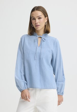 Woman wearing a light blue long-sleeve blouse with a tie neckline and white pants, standing against a plain white background.