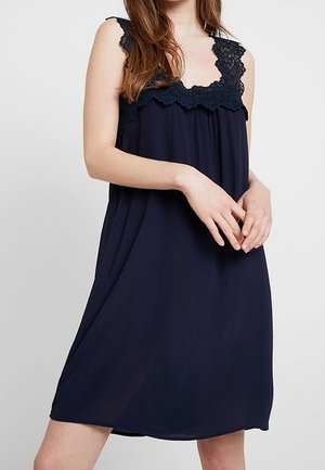 Woman wearing sleeveless navy blue dress with lace trim at the neckline, standing with hands loosely clasped in front.