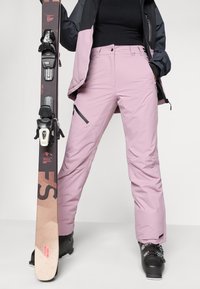 Pantaloni de schi de un roz deschis, cu o croială lejeră, buzunare cu fermoar și tivuri întărite. Inclusiv boote de schi negre și o skie cu legături.
