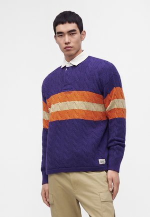 Ung mand iført en lilla kabelstrikket sweater med orange og beige striber over en hvid skjorte med krave og beige bukser.