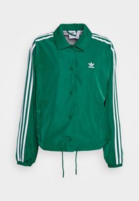 Chaqueta de nailon verde con rayas blancas en las mangas, botones a presión, puños elásticos y dobladillo con cordón. Presenta un pequeño logo en el pecho.