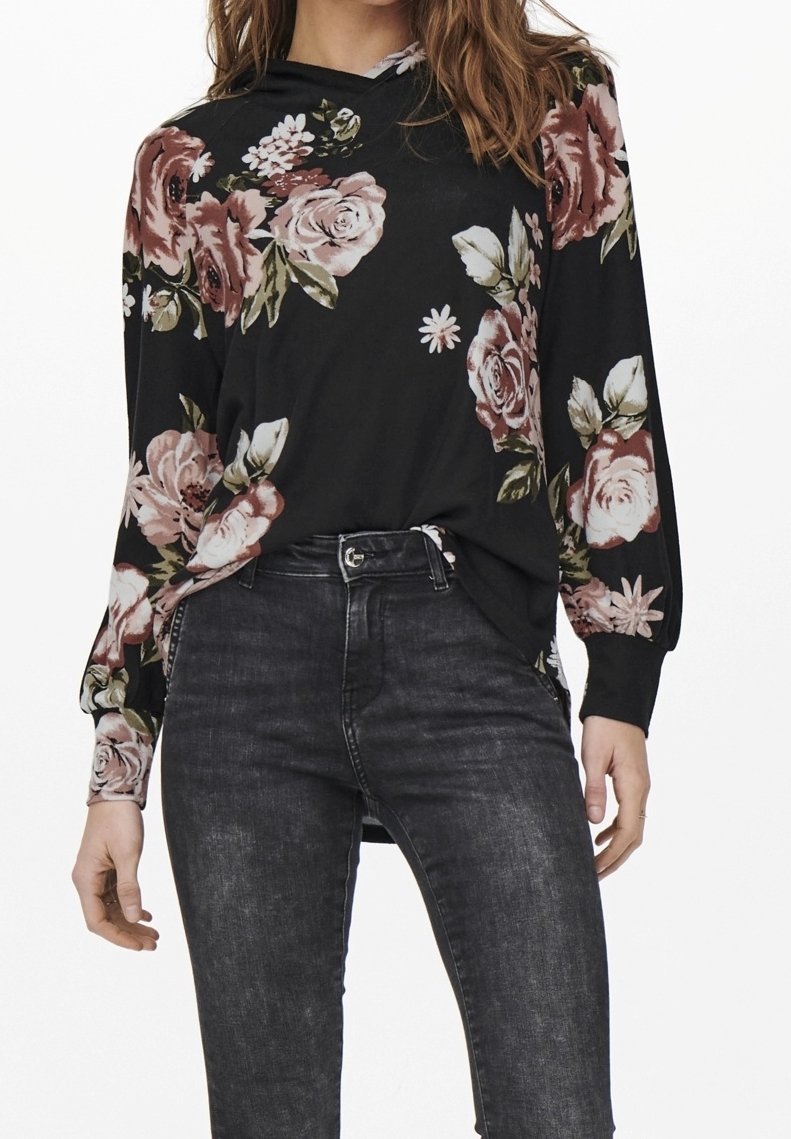 Femme portant un chemisier noir à manches longues avec un imprimé floral rose et blanc, rentré dans un jean noir délavé taille haute.