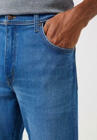 Niebieskie dżinsy o klasycznym kroju, z przednią kieszenią, widocznym przeszyciem i delikatnym połyskiem na tkaninie.