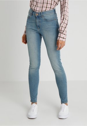 Mujer con jeans ajustados azul claro, zapatillas blancas y una camisa de cuadros rosa y blanca de manga larga, de pie frente a un fondo liso.