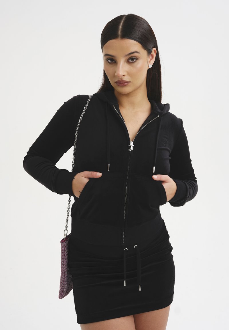 Felpa nera con zip, dal design aderente, texture morbida e tasca a marsupio frontale. Abbinata a una mini gonna nera arricciata e una borsa a catena metallica.