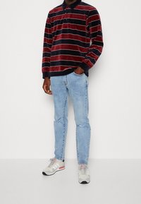 Man in een rood en zwart gestreepte lange mouwen polo, lichtblauwe ripped jeans en witte sneakers, staand tegen een witte achtergrond.