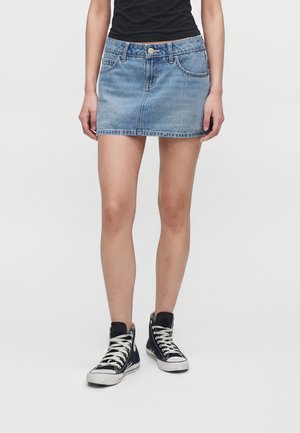 Person iført en lyseblå denim mininederdel, sorte høje sneakers med hvide snørebånd og en sort top mod en ensfarvet baggrund.