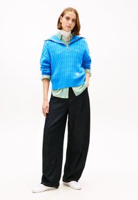 Pulover albastru cu model tricotat în cablu și guler cu fermoar, purtat peste o cămașă verde cu dungi, asortat cu blugi largi și întunecați și pantofi albi.