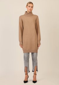 Beige gestrickter Rollkragensweater mit übergroßen Ärmeln, kombiniert mit hellgrauen Jeans mit Fransen an den Säumen und mit Perlen verziert. Schwarze Absatzschuhe.