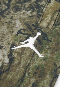 Tkanina s camouflage vzorom v odtieňoch zelenej a hnedej obsahuje bielu vyšívanú logo Jumpman a subtílne čierne textové akcenty.