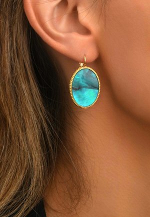 Gros plan d’une boucle d’oreille créole en or avec une grande pierre turquoise ovale portée sur l’oreille d’une personne aux cheveux châtain clair.