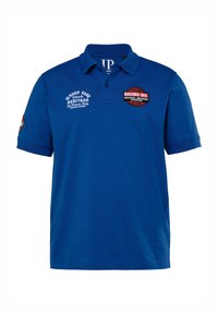 Modrá bavlněná polo košile s límečkem. Obsahuje výšivku na hrudi a nášivky, včetně "RACING DIV" a "CLASSIC CARS HERITAGE."