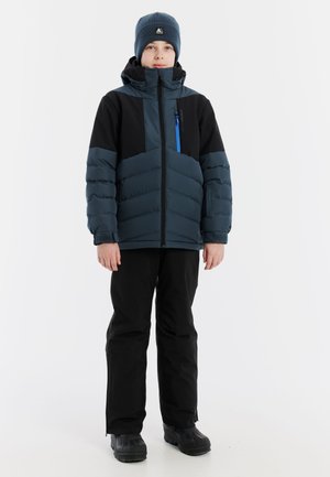 Blauer Puffer-Jacke mit schwarzen Akzenten, mit Reißverschlusstasche, kombiniert mit schwarzen Hosen und schwarzen Stiefeln. Modell trägt eine blaue Mütze.