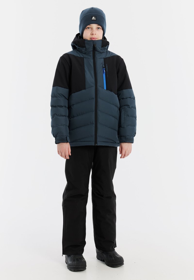 Blauer Puffer-Jacke mit schwarzen Akzenten, mit Reißverschlusstasche, kombiniert mit schwarzen Hosen und schwarzen Stiefeln. Modell trägt eine blaue Mütze.