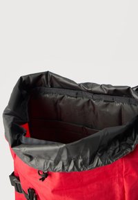 Κόκκινο σακίδιο με μαύρο κορδόνι στο άνοιγμα, που αποκαλύπτει έναν ευρύχωρο εσωτερικό χώρο επενδεδυμένο με μαύρο ύφασμα και εσωτερικές τσέπες οργάνωσης.