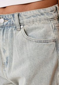 Jeans de denim azul claro con cintura alta, que presentan remaches clásicos, un bolsillo delantero y un sutil desgaste en la textura de la tela.