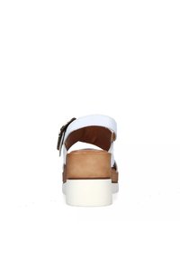 Sandal bianca con una fascetta ampia e tacco, con un accento marrone. Hardware con fibbia regolabile e suola testurizzata. Sfondo neutro.