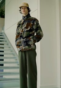 Fleecejakke i camouflage-mønster med grønne, brune og blå nuancer, parret med strukturerede grønne bukser og en tan cap.