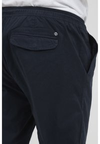 Close-up van marineblauwe casual broek met elastische tailleband, een klep achterzak en zichtbare zijzak, gedragen door een persoon in een witte shirt.