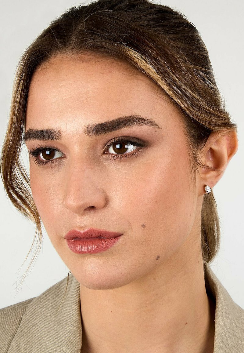 Närbild av en models ansikte med medelhudton, ljust brunt hår, bärande små, runda diamantörhängen och neutralläppstift.