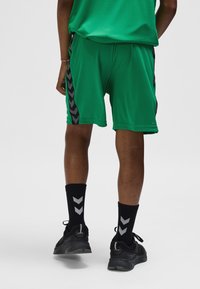 Groene sportshorts met zwarte chevron-zijstrepen, gecombineerd met zwarte sokken met grijze chevron-accenten en zwarte sneakers.