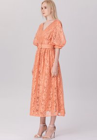 Robe midi en dentelle corail avec un décolleté en V, des manches bouffantes, une taille ceinturée et des motifs floraux élaborés. Possède une silhouette fluide.