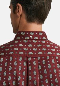 Homme aux cheveux courts et foncés portant une chemise rouge avec des motifs paisley et circulaires bleus et jaunes, vu de dos.