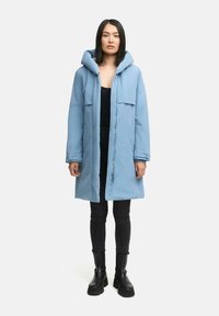 Parka azul claro con capucha, cremallera frontal y dos bolsillos en el pecho, usada sobre una blusa negra y pantalones negros, combinada con botas negras.