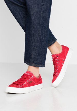 Benen met donkerblauwe spijkerbroek en rode canvas sneakers met witte zolen, halverwege een stap op een lichtgrijze ondergrond en achtergrond.