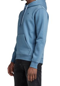 Hoodie bleu clair avec capuche à cordon, poche kangourou, poignets et ourlet côtelés, portant un logo discret sur la poitrine. Tissu doux au toucher.