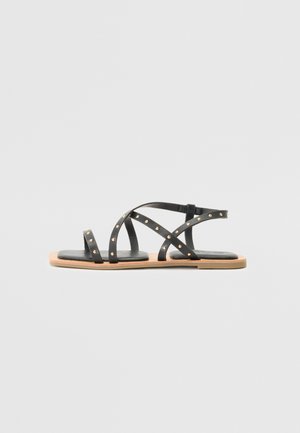 Black leather sandals with multiple crisscross straps, embellished with gold studs, and a flat tan sole. Side view displayed.