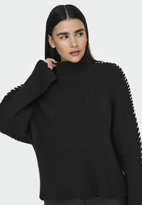 Maglione nero a coste con un collo alto e maniche ampie. Presenta decorativi tagli lungo le braccia e una vestibilità morbida. Tessuto morbido.