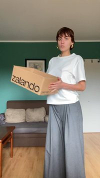 Caixa de cartão com "zalando" impresso em letras grandes, segurada por uma pessoa vestindo uma t-shirt branca e calças largas cinzas, em um ambiente interior.