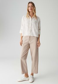 Camicia bianca a bottoni con texture e maniche a tre quarti, abbinata a pantaloni beige chiaro a gamba larga e scarpe da ginnastica bianche.