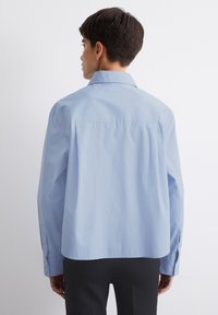 Lysblå skjorte med knapper lavet af stof med en glat tekstur. Har en spidse krave og lange ærmer, med en løs pasform og rygslids.