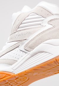 Gros plan d'une basket en daim blanc et beige avec cuir perforé, détails côtelés, broderie signature et semelle en caoutchouc orange.