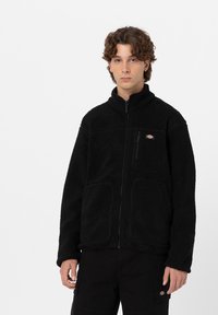 Sort fleece jakke med høj krave, frontlynlås og to store sidelommer. Har et lille logo på brystet og tekstureret stof.