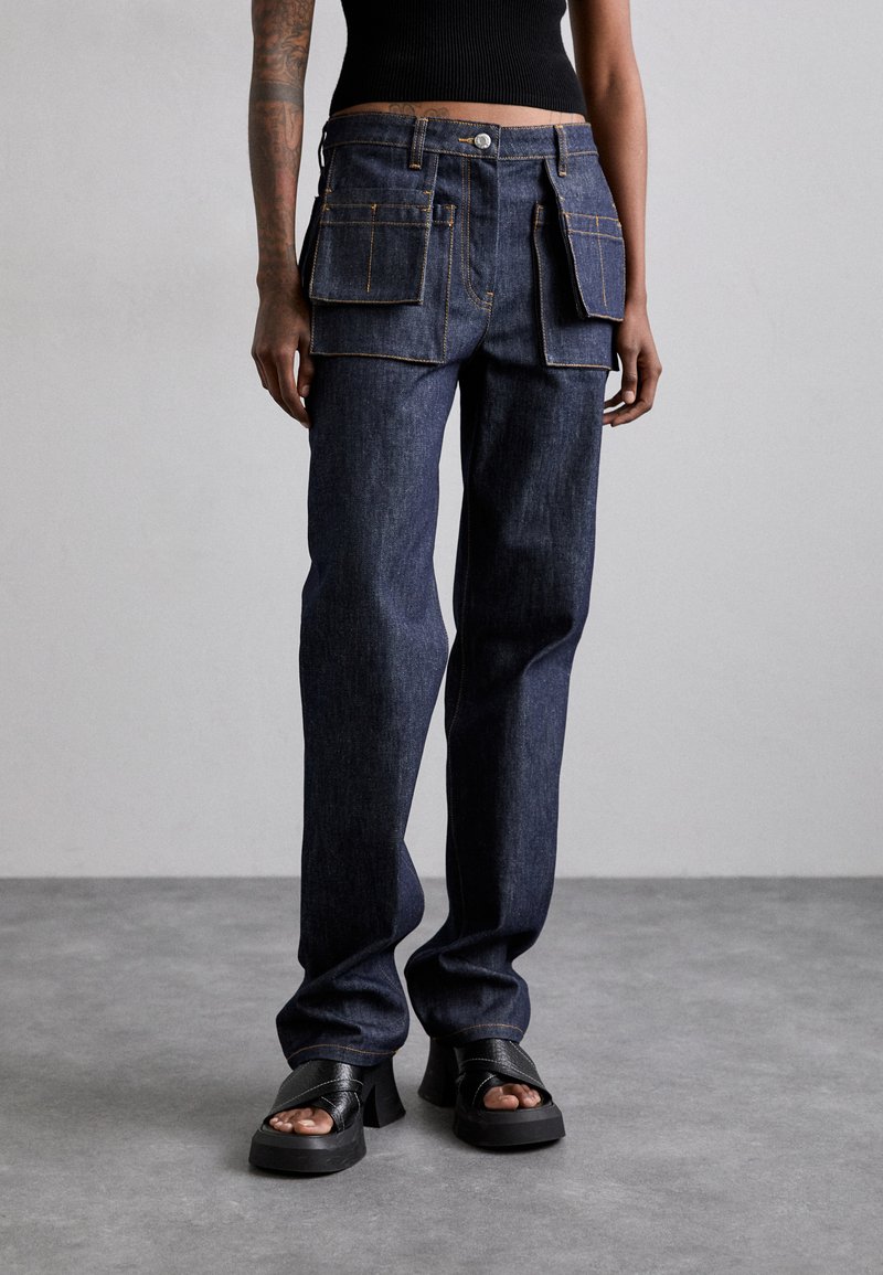 Pantaloni cargo din denim albastru închis, cu două buzunare mari în față și cusături portocalii. Design cu picior drept, purtați cu sandale negre cu toc pătrat.
