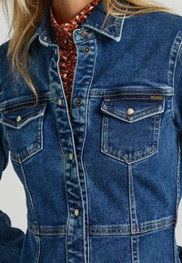 Blå denimjacka med en strukturerad design, som har två bröstfickor, metallknappar och synliga söm detaljer.