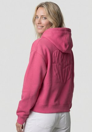 Blonde Frau lächelt über die Schulter, trägt einen leuchtend pinken Hoodie mit erhabenen Schriftzügen auf der Rückseite und weiße Hosen vor einem schlichten Hintergrund.