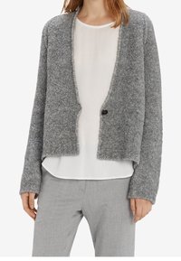 Šedý, měkký pletený kardigan s volným střihem, otevřeným předním dílem a jedním černým knoflíkem. Kombinovaný s bílou blůzou a šedými kalhotami.