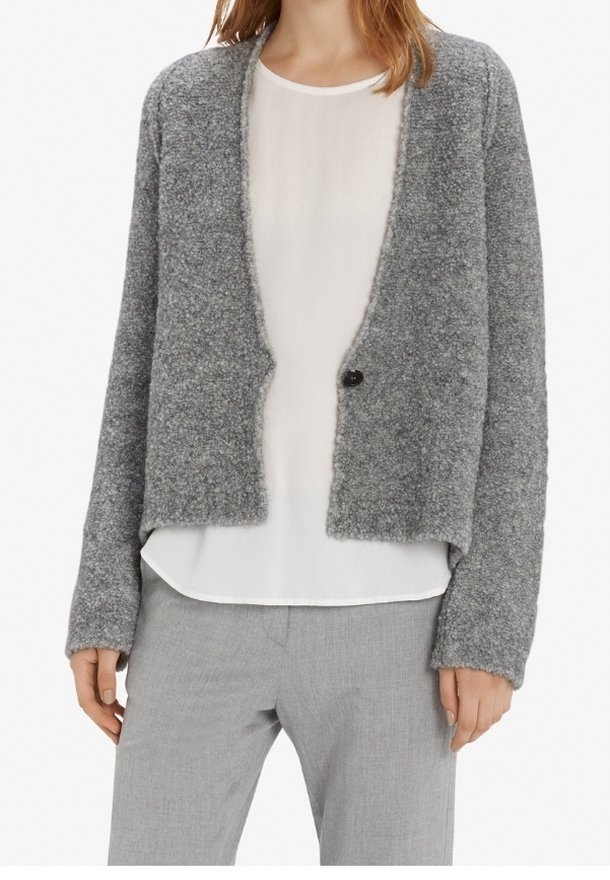 Šedý, měkký pletený kardigan s volným střihem, otevřeným předním dílem a jedním černým knoflíkem. Kombinovaný s bílou blůzou a šedými kalhotami.
