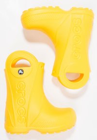 Bottes de pluie en caoutchouc jaunes avec une surface texturée, une poignée découpée en cercle et un logo "CROCS" en relief sur le côté. Design léger et étanche.