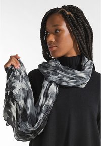 Sciarpa leggera e texture in tonalità di nero e grigio con un design ondulato, appoggiata casualmente sulle spalle e tenuta in una mano.