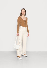 Blusa longa castanha com decote em V e fecho de laço, combinada com calças largas brancas de cintura alta. Uma pequena bolsa branca com alça studada.