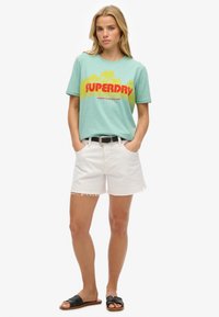 Mint grøn t-shirt med "SUPERDRY" grafik i rød og gul, parret med hvide denim shorts med frynset kant og sorte slidesandaler.